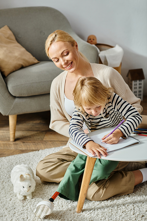 blonde girl with prosthetic leg drawing on paper with colorful pencils near happy mother at home