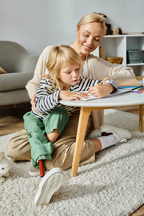 happy blonde woman looking at her daughter with prosthetic leg drawing on paper with colorful pencil