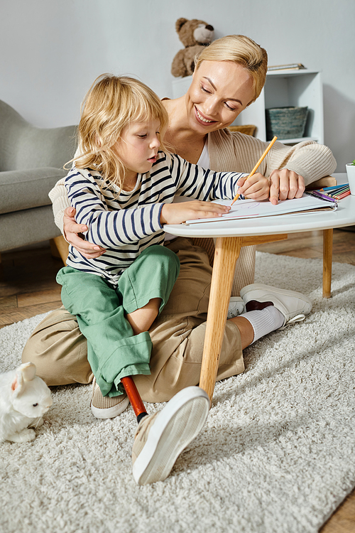 happy mother looking at her daughter with prosthetic leg drawing on paper with colorful pencil