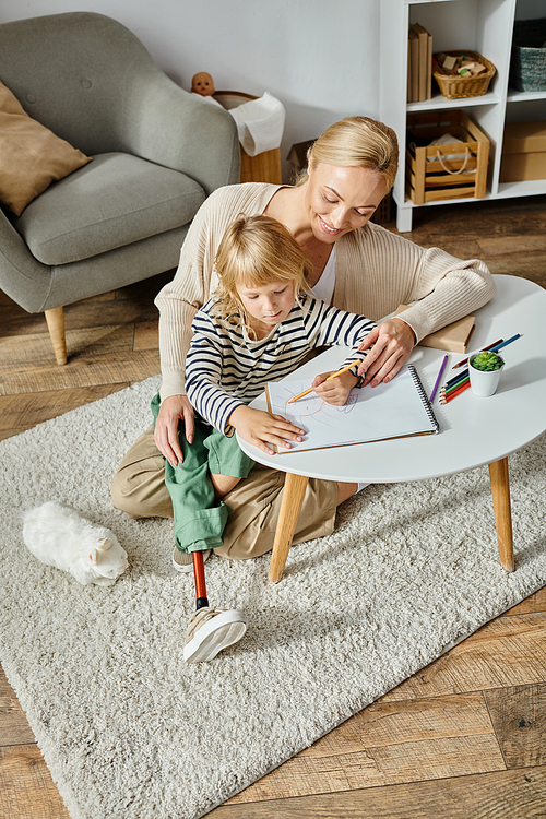 happy blonde woman looking at her daughter with prosthetic leg drawing on paper with colorful pencil