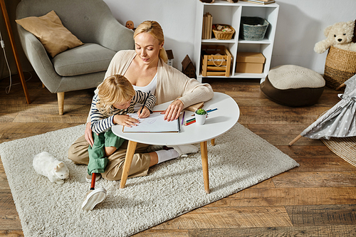 happy woman looking at daughter with prosthetic leg drawing on paper with colorful pencil, overhead