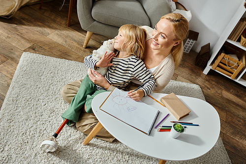 happy woman hugging daughter with prosthetic leg near paper and colorful pencil on table, overhead