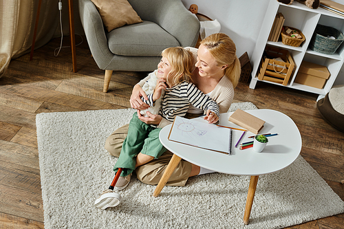 woman hugging happy daughter with prosthetic leg near paper and colorful pencil on table, overhead