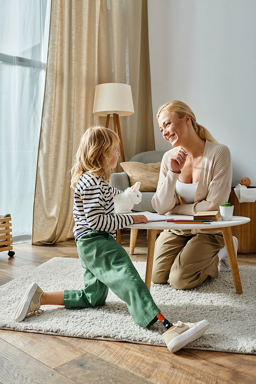 happy mother looking at daughter with prosthetic leg sitting near table with drawing and soft toy