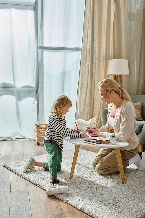 happy mother looking at her daughter with prosthetic leg drawing on paper with colorful pencil