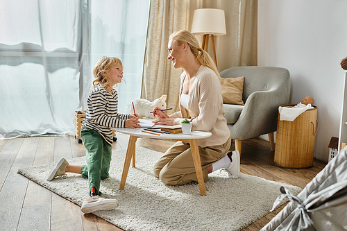 happy mother looking at her kid with prosthetic leg drawing on paper with colorful pencil