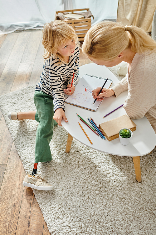 mother and kid with prosthetic leg drawing together on paper with colorful pencils, quality time