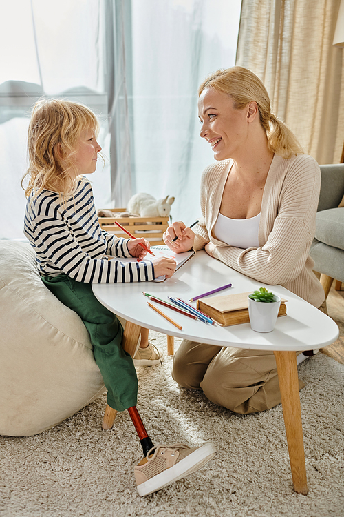 mother and kid with prosthetic leg drawing on paper with colorful pencils together, quality time