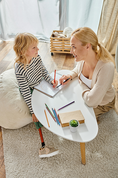 happy mother and kid with prosthetic leg drawing on paper with colorful pencils, quality time