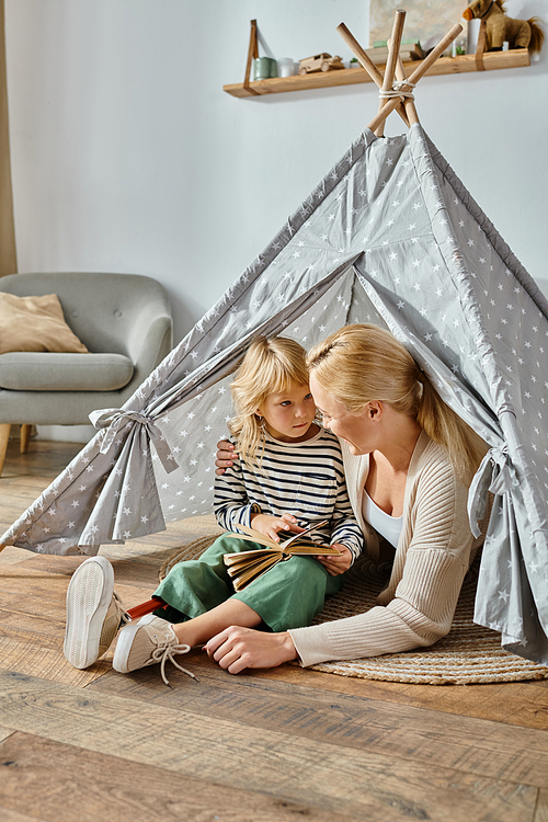 cute girl with prosthetic leg reading book and sitting with happy mother in play tent at home