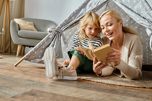 cute girl with prosthetic leg and blonde mother reading book and sitting in play tent at home