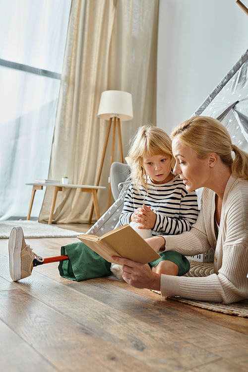 cute girl with prosthetic leg holding soft toy and listening to mother reading book in play tent