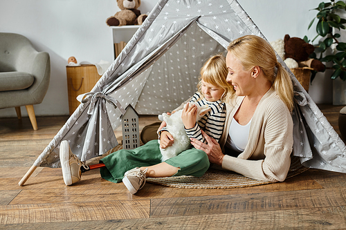 little girl with prosthetic leg hugging soft toy rabbit near happy mother while sitting in play tent