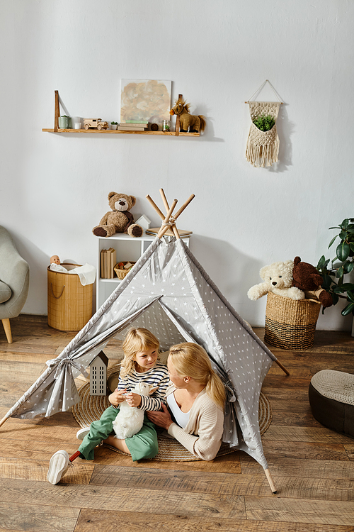 disabled girl with prosthetic leg holding toy rabbit near happy mother while sitting in play tent