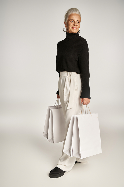 happy middle aged woman in elegant attire and hoop earrings standing with shopping bags on grey
