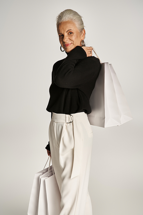 cheerful middle aged woman in elegant attire and hoop earrings standing with shopping bags on grey