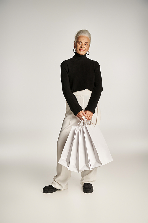 satisfied middle aged woman in elegant attire and hoop earrings standing with shopping bags on grey