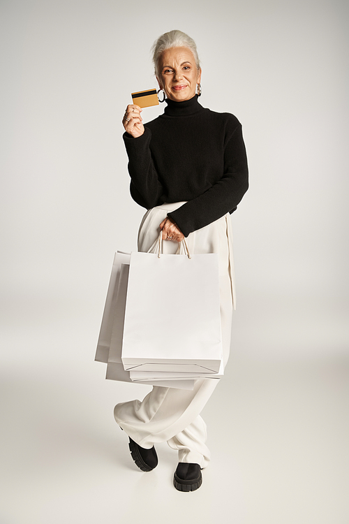 satisfied middle aged woman in elegant attire holding credit card and shopping bags on grey backdrop