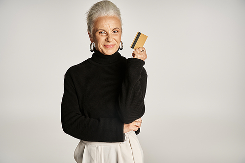 elegant middle aged woman in black turtleneck sweater holding credit card on grey backdrop