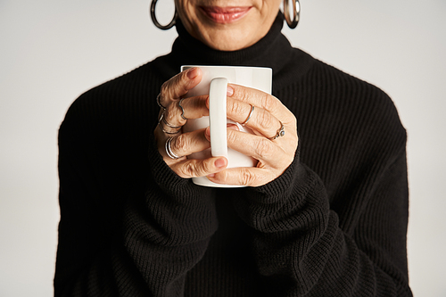 cropped middle aged woman in black turtleneck sweater holding cup with hot beverage on grey backdrop