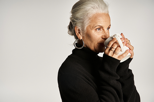 happy middle aged woman in black turtleneck sweater holding cup of coffee on grey backdrop