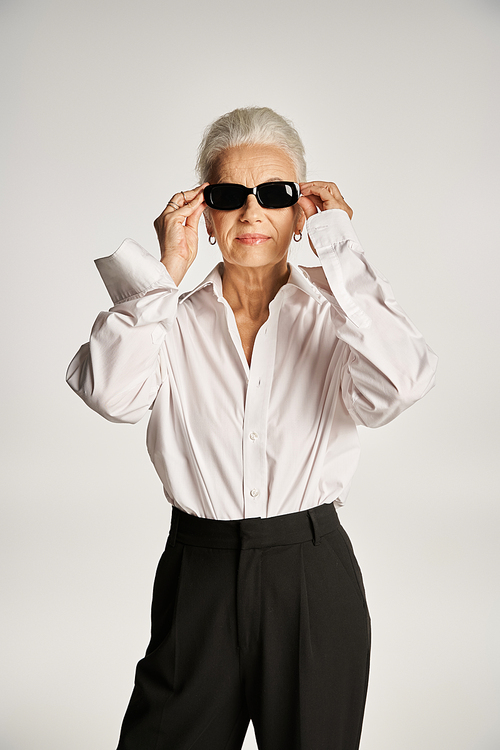 stylish middle aged woman with grey hair wearing sunglasses and standing in elegant attire on grey