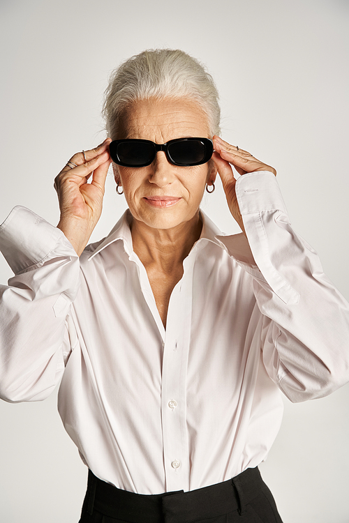 stylish middle aged woman with grey hair wearing sunglasses and standing in elegant attire on grey