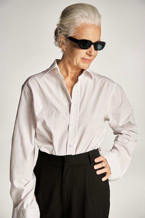 middle aged woman in sunglasses and elegant attire standing with hand on hip on grey backdrop