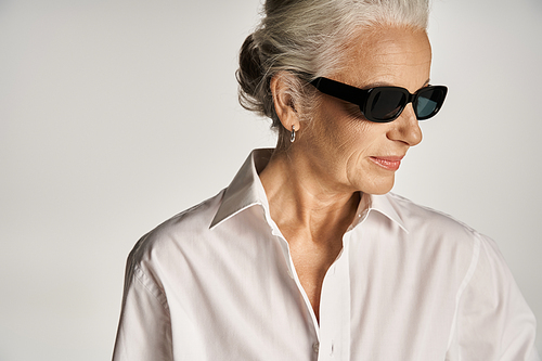 portrait of stylish middle aged woman in white shirt and sunglasses posing on grey background