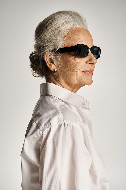portrait of stylish middle aged woman in white shirt and sunglasses posing on grey background