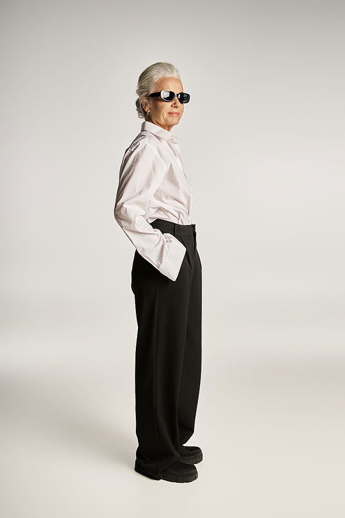 fashionable middle aged woman in white shirt and sunglasses posing with hand in pocket on grey