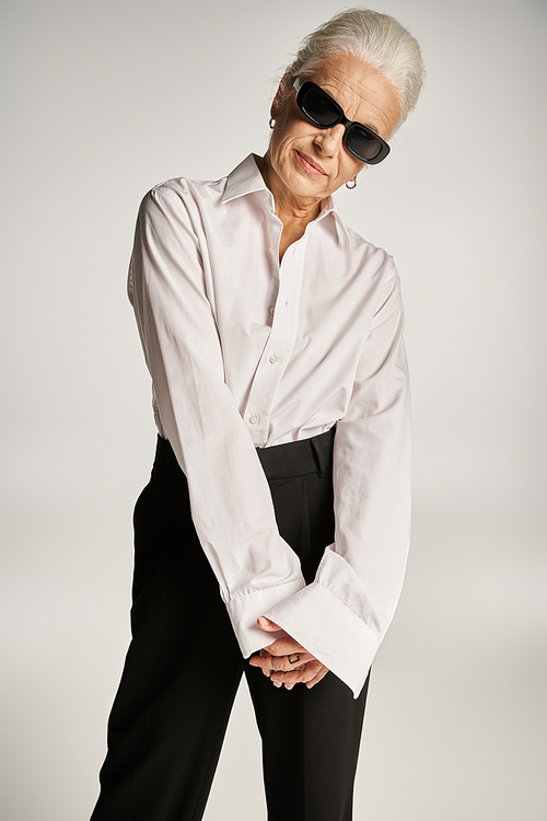 fashionable middle aged woman in white shirt, black pants and sunglasses posing on grey backdrop