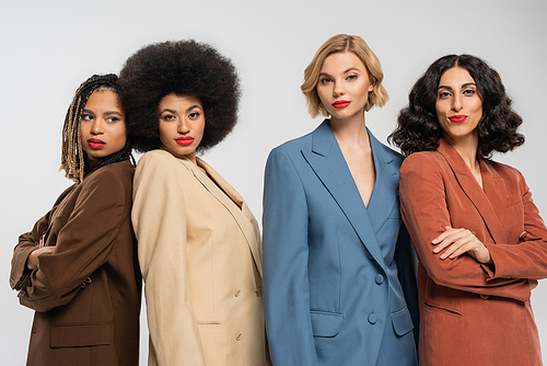 group of elegant multiethnic women in colorful suits looking at camera on grey, trendy girlfriends