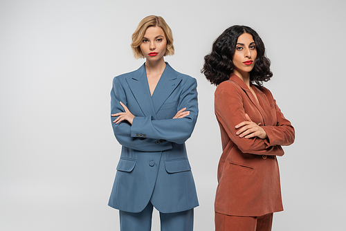 multiethnic blonde and brunette female models in colorful suits posing with folded arms on grey