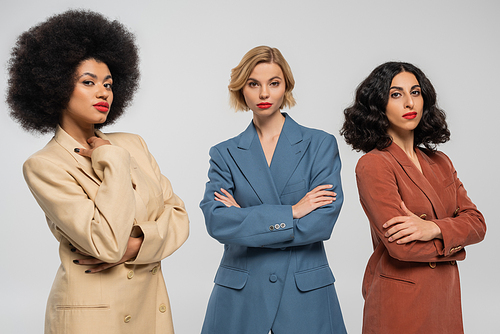 three confident multiracial girlfriends in multicolored suits standing with folded arms on grey