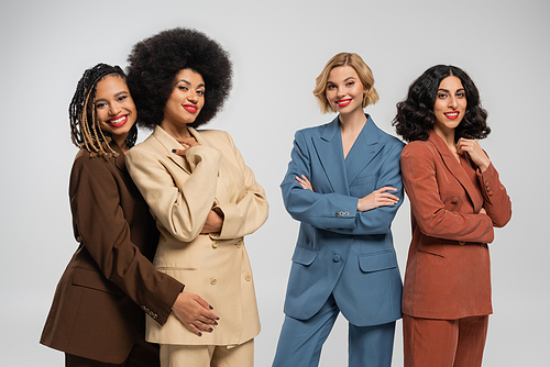 group of trendy multiethnic girlfriends in multicolored suits smiling at camera on grey, diversity