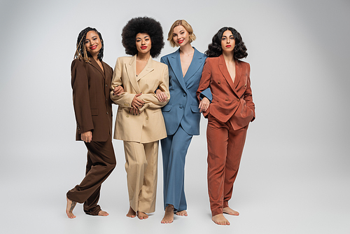 multicultural barefoot female models standing in colorful suits on grey backdrop, full length
