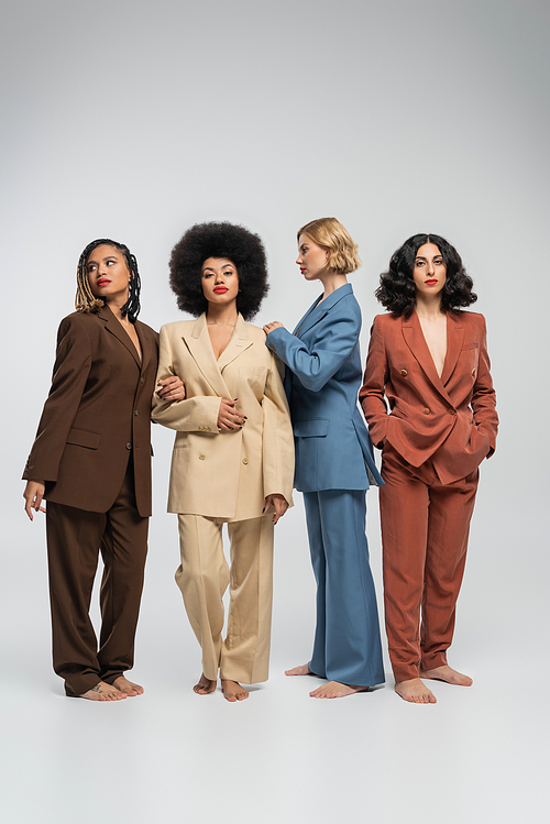 multicultural barefoot women in multicolored suits standing on grey backdrop, fashion photography