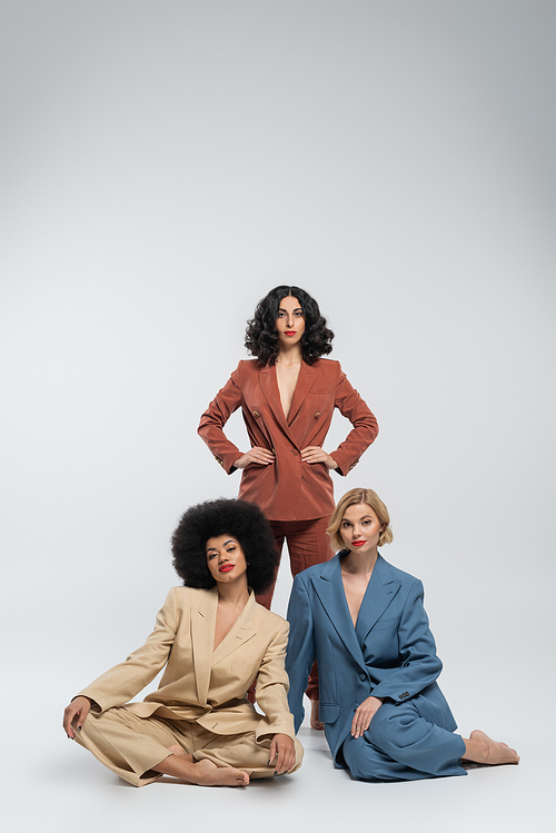 stylish multiracial woman with hands on hips near girlfriends in colorful suits sitting on grey