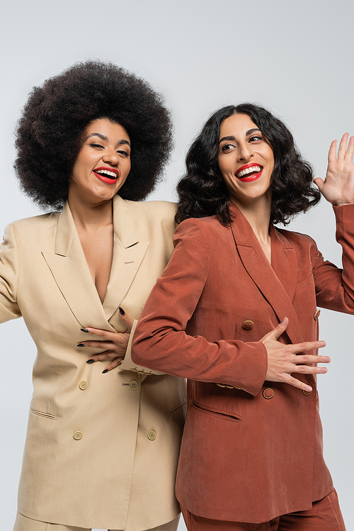 cheerful multiracial woman waving hand near african american girlfriend on grey, colorful suits