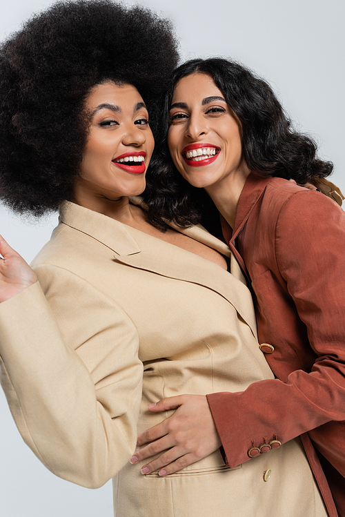 happy multiracial woman embracing african american girlfriend on grey, models in colorful suits