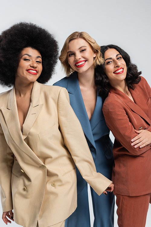 smiley multiethnic girlfriends in multicolored suits looking at camera on grey, diverse beauty