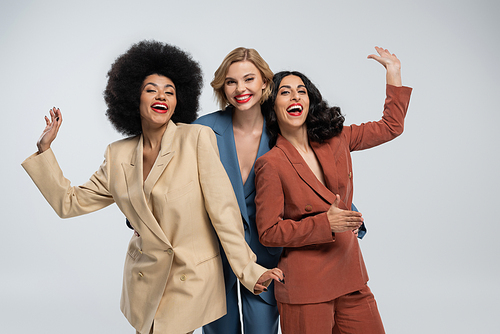 joyful multiethnic girlfriends in trendy colorful suits waving hands at camera on grey, modern style