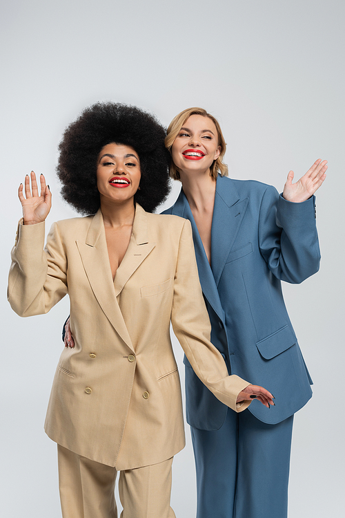 smiling multiethnic female friends in multicolored suits waving hands on grey, fashion shoot