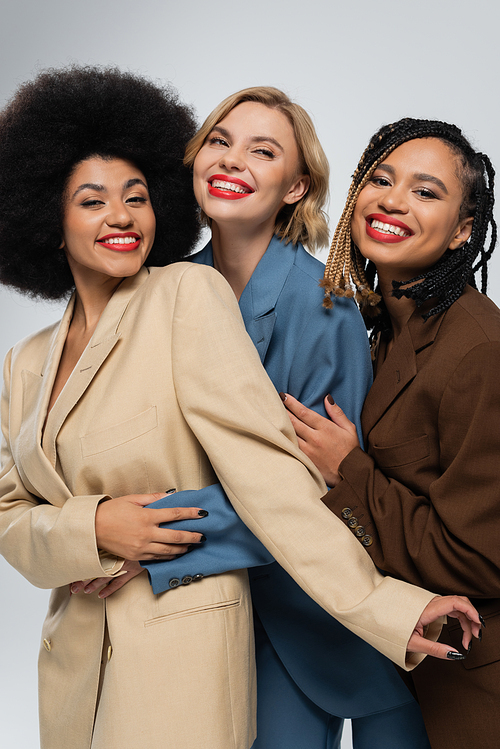 happy multiracial girlfriends in multicolored suits embracing on grey, togetherness and style