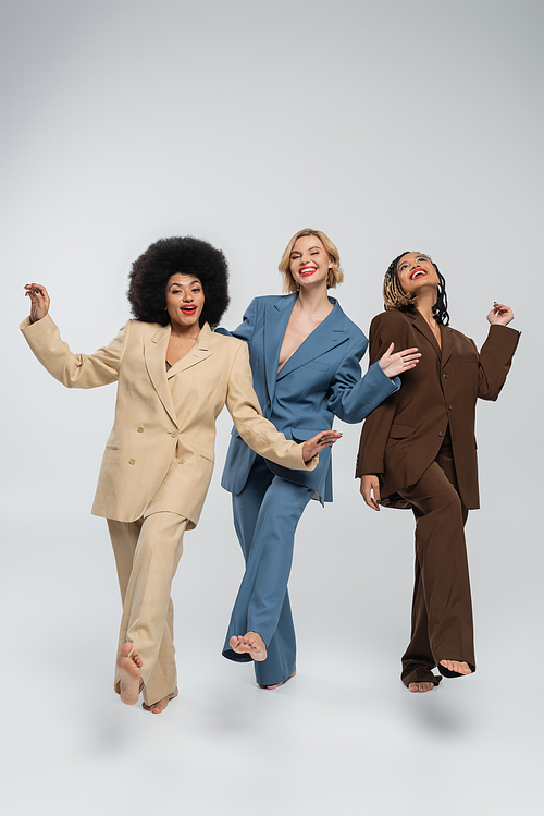 joyful barefoot multiracial fashion models in colorful suits posing barefoot on grey, full length