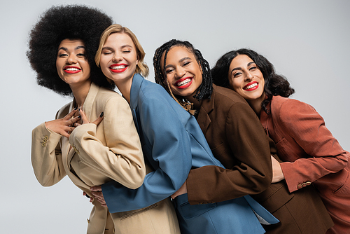 group of trendy multiracial girlfriends in colorful suits embracing and smiling at camera on grey