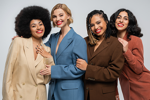 four jolly multiracial girlfriends in trendy colorful suits smiling at camera on grey, diversity