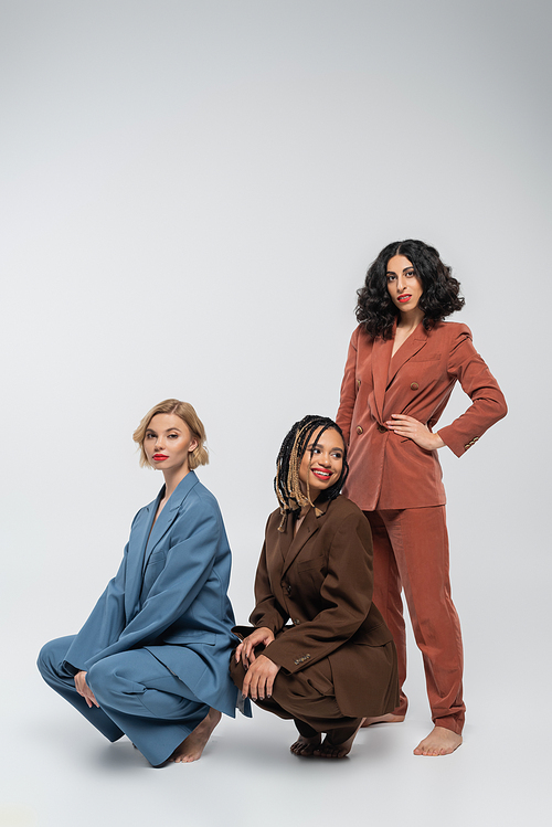 multiracial woman with hand on hip near girlfriends in colorful suits sitting on haunches on grey