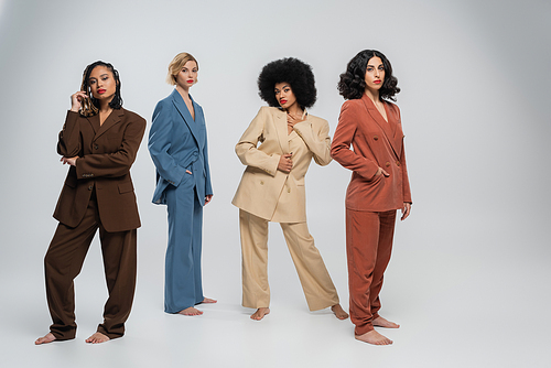 full length of multiracial barefoot female friends in trendy multicolored suits standing on grey